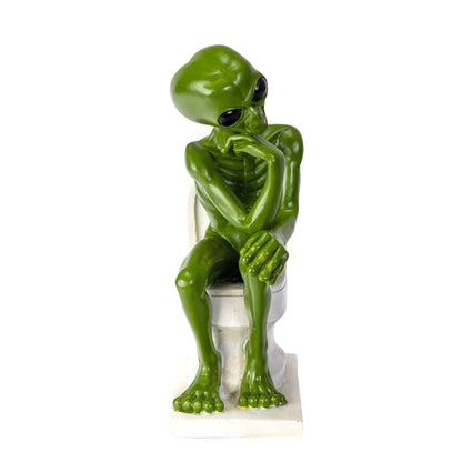 Front view of alien figurine sitting on toilet with hands resting
