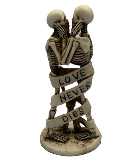 Front view of gothic skeleton couple statue standing and embracing with “Love Never Dies” ribbon wrapped around bodies
