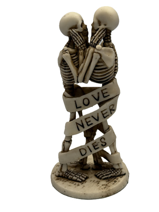 Front view of gothic skeleton couple statue standing and embracing with “Love Never Dies” ribbon wrapped around bodies
